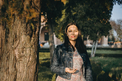 Portrait of smiling young woman standing by tree trunk