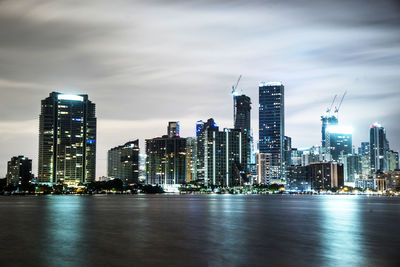 Illuminated city by sea against cloudy sky at night