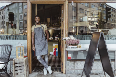 Smiling male owner leaning on doorway of delicatessen shop