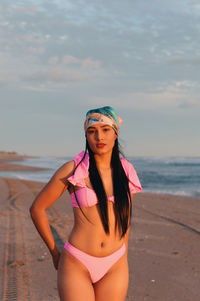 Portrait of young woman standing at beach against sky during sunset