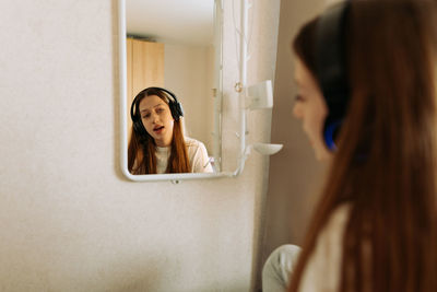 Beautiful teenage girl in headphones singing at the mirror at home