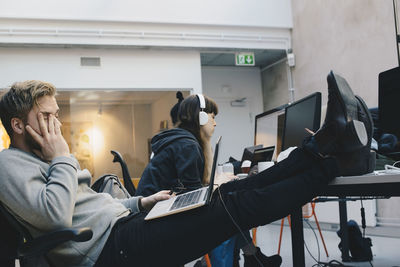 Tired computer programmer sitting with feet up on desk while colleagues working in background