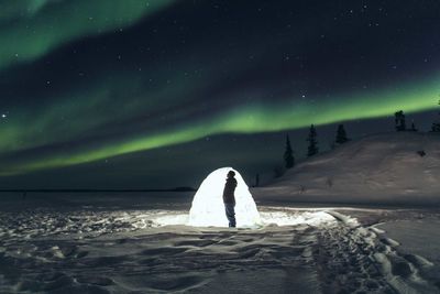 Man standing by illuminated tent on snow covered field at night