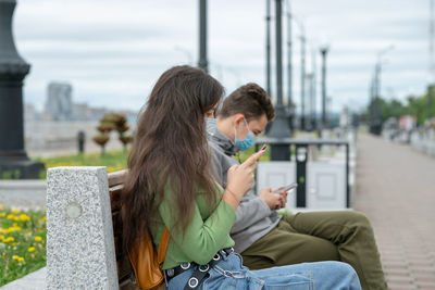 A guy and a girl are sitting on a bench at a distance from each other with masks on their faces 