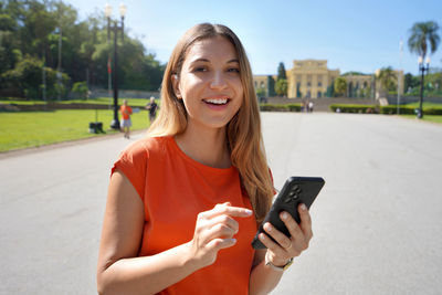 Young woman looking at camera and holding mobile phone outdoor with blurred city park on background