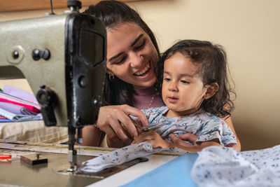 Cheerful hispanic female looking at cute cute girl with smile while sitting at professional sewing machine during work in light workshop