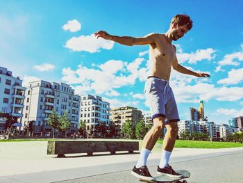 Full length of shirtless man skateboarding on street by buildings against sky