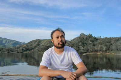 Man looking away while sitting by lake against sky