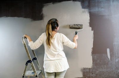 Woman climbed a ladder and catching a paint roller full of grey painting on a brown wall. painter is upping and downing roller covering the wall with grey painting what remains wet. horizonta