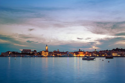 Illuminated buildings at waterfront against cloudy sky