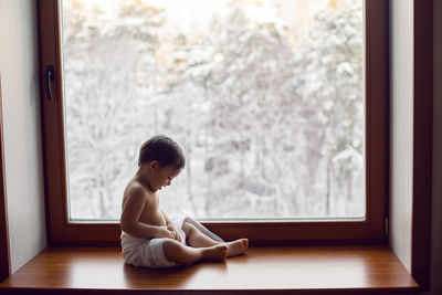 Baby boy in bath towel after washing sitting on the windowsill at the big window in winter