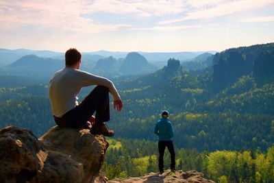 Boy and girl tourists stay on cliff and thinking. wild nature, blue misty sunrise in valley below