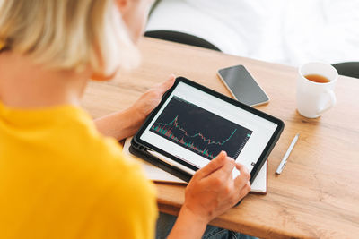 Young blonde woman check cryptocurrency charts and stock quotes on digital tablet with cup at home