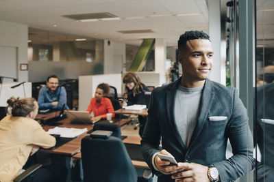 Mid adult businessman holding smart phone looking through window at office