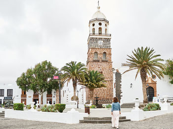 Unrecognizable woman walking towards fountain and tropical trees on town square near brick tower on gray day in fuerteventura, spain