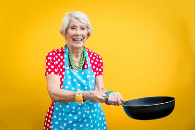 Portrait of smiling senior woman holding cooking pan against yellow background