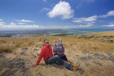 Couple sitting on mountain against blue sky during sunny day