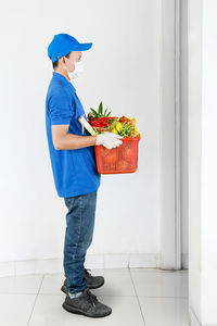 Full length of delivery person wearing mask holding fruits standing outdoors