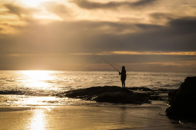 Silhouette person fishing at sea against cloudy sky during sunset