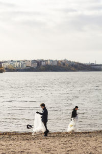 Young male environmentalists collecting plastic waste by lake against clear sky