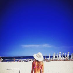 Rear view of woman in bikini wearing sun hat at beach against blue sky