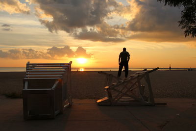 Rear view of silhouette man standing on fallen lifeguard chair at beach during sunset