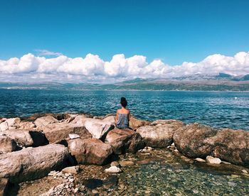 Rear view of woman sitting in rock by sea against blue sky