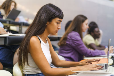 Side view of young female student using laptop while sitting in lecture hall at university