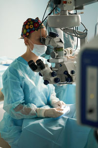 Adult female doctor in sterile mask and ornamental medical cap looking through surgical microscope against crop coworker in hospital