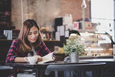 Smiling young woman reading book while sitting by coffee on table at cafe