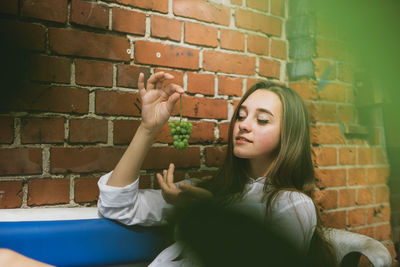 Smiling woman holding grapes while sitting by brick wall