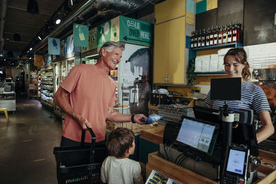 Happy senior man talking to cashier while standing with grandson at checkout counter in supermarket