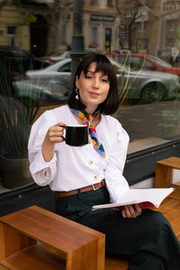 Portrait of young woman holding book and coffee cup while sitting outdoors