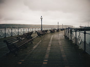 Empty benches on pier over sea against cloudy sky