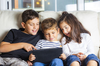Happy siblings using digital tablet while sitting on sofa at home