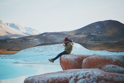 Side view of man photographing while sitting on rock against sky