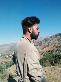 Side view of young man looking away while standing on mountain against clear blue sky