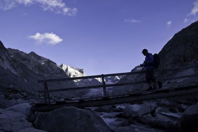 Man crossing stream on footbridge by mountain against sky