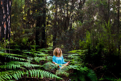 Woman with curly hair meditating while sitting amidst plants in forest