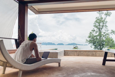 Rear view of woman sitting on chair by swimming pool against window