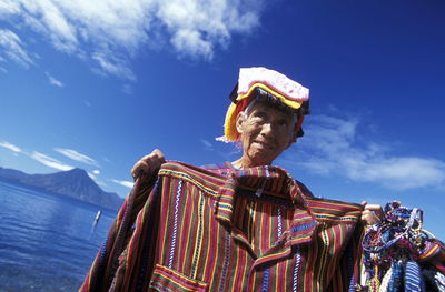 Low angle view of senior woman selling shirt by lake against sky