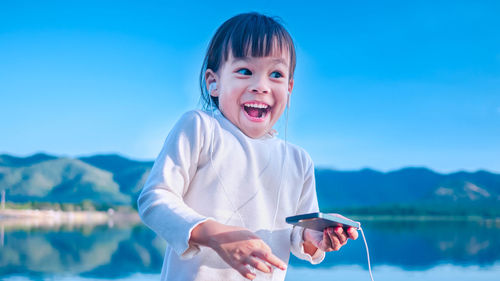 Cheerful girl listening music while standing against lake