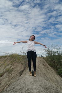 Happy woman with arms outstretched standing on rock against sky