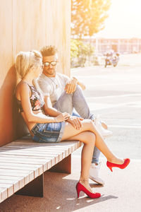 Mid adult couple sitting on boardwalk against wooden wall during sunny day