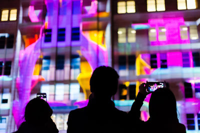 Rear view of silhouette people photographing illuminated building at night