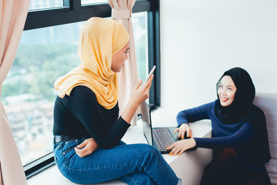 Young woman using mobile phone while sitting on window