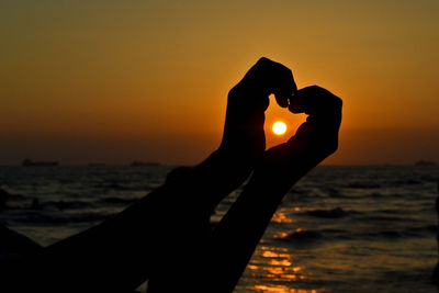 Close-up of silhouette hand making heart shape against orange sky during sunset