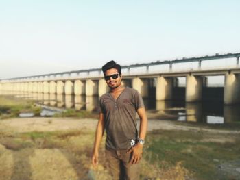 Young man wearing sunglasses standing against bridge against clear sky
