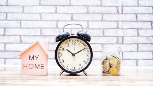 Close-up of alarm clock by coins and model home on table against wall