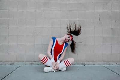 Woman in sports costume tossing hair while sitting on sidewalk against wall